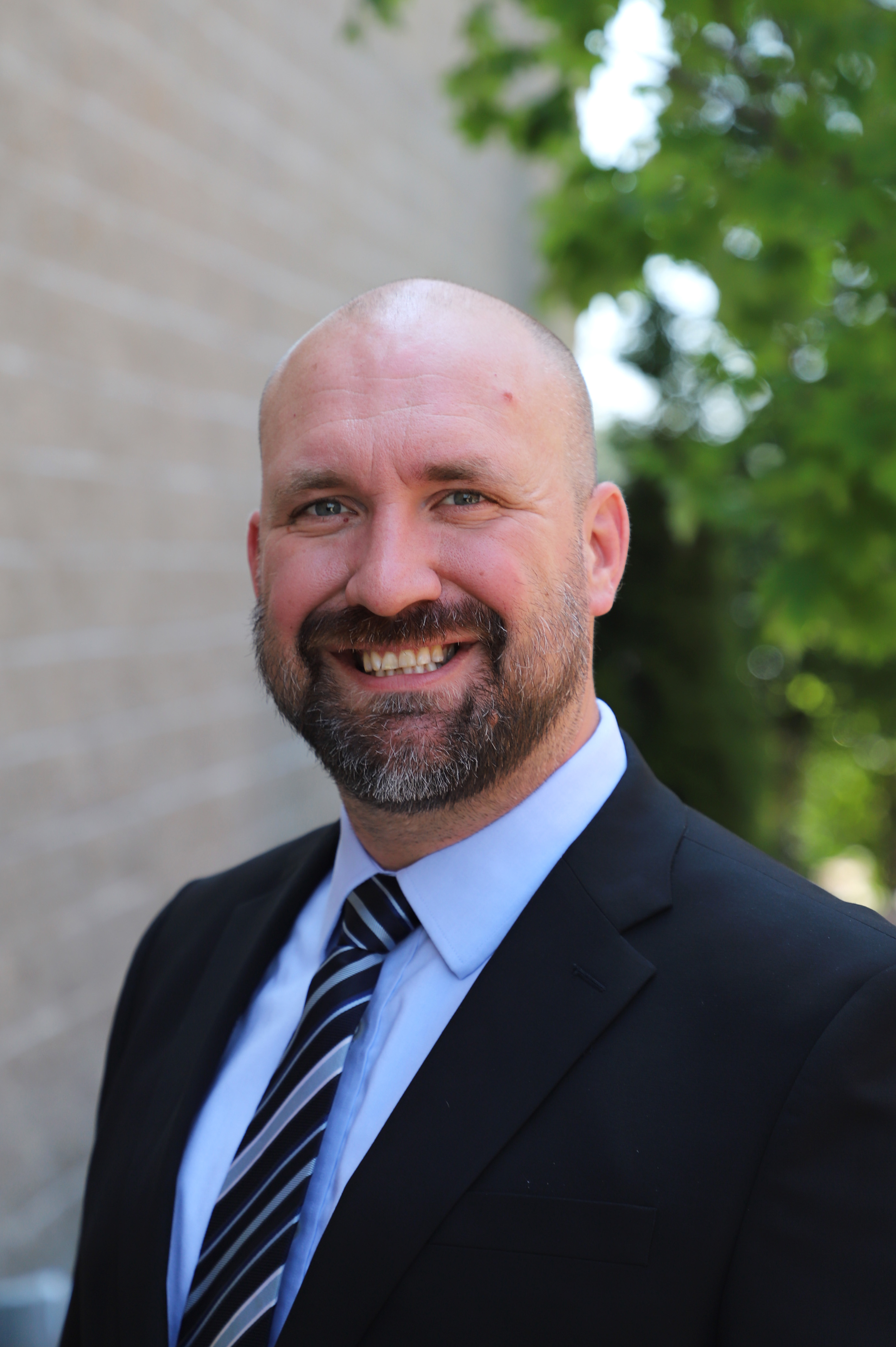 Headshot of Matt Rodda outdoors, blue shirt and tie with black jacket, green trees and brick wall behind.